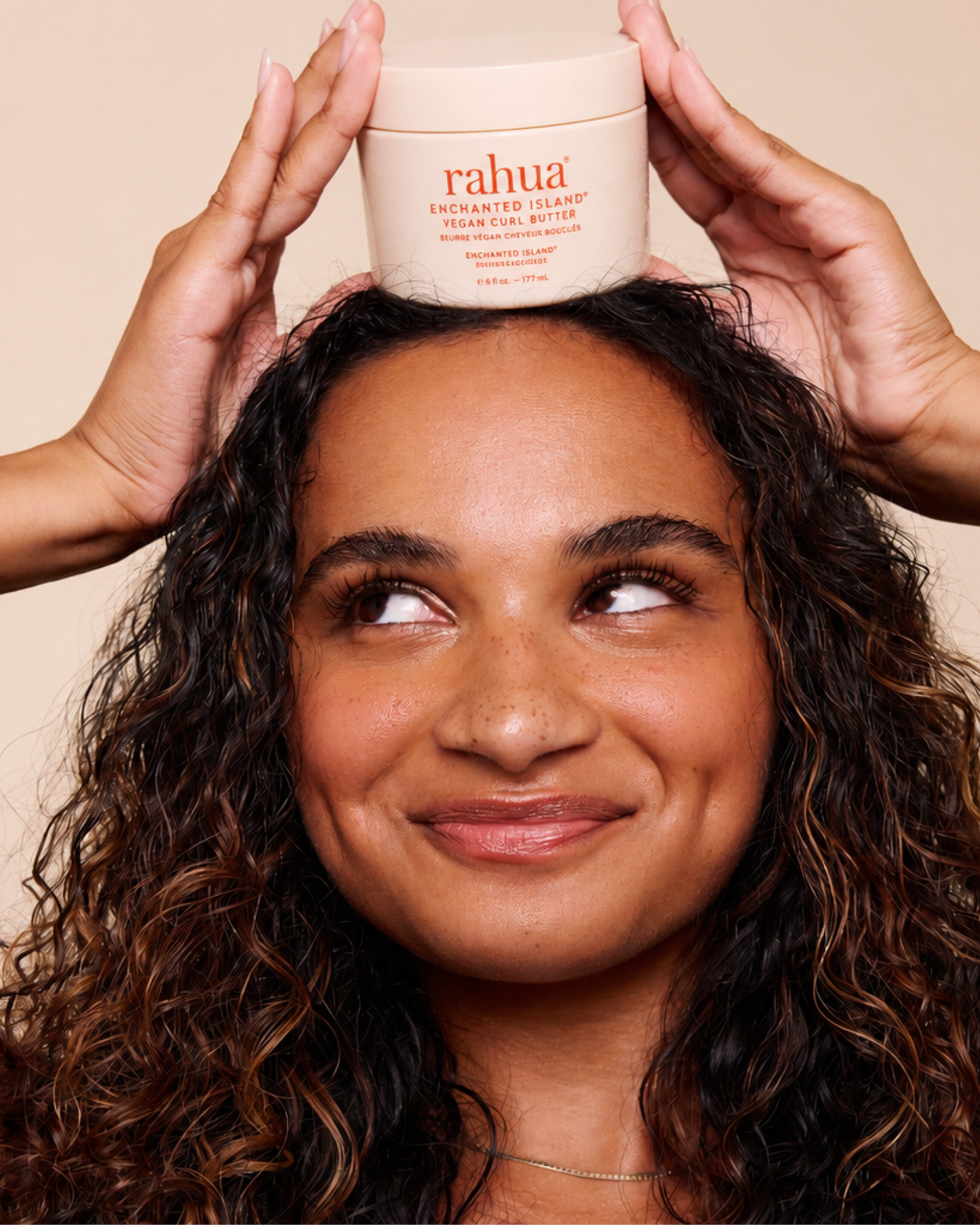 Woman holding a jar of rahua hair product on her head against a beige background